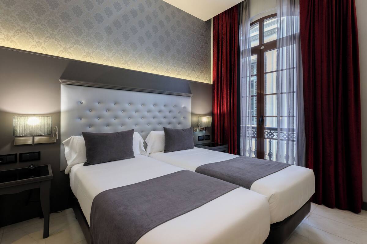 Habitación de hotel con dos camas, un cabecero grande, una mesita de noche con lámpara y una ventana con cortinas rojas.