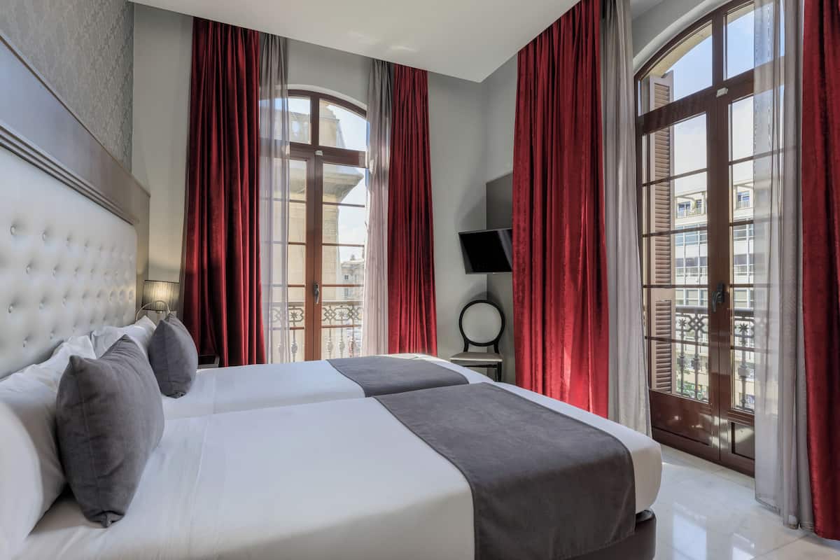 Habitación de hotel con dos camas, televisor y ventanas amplias con cortinas rojas.