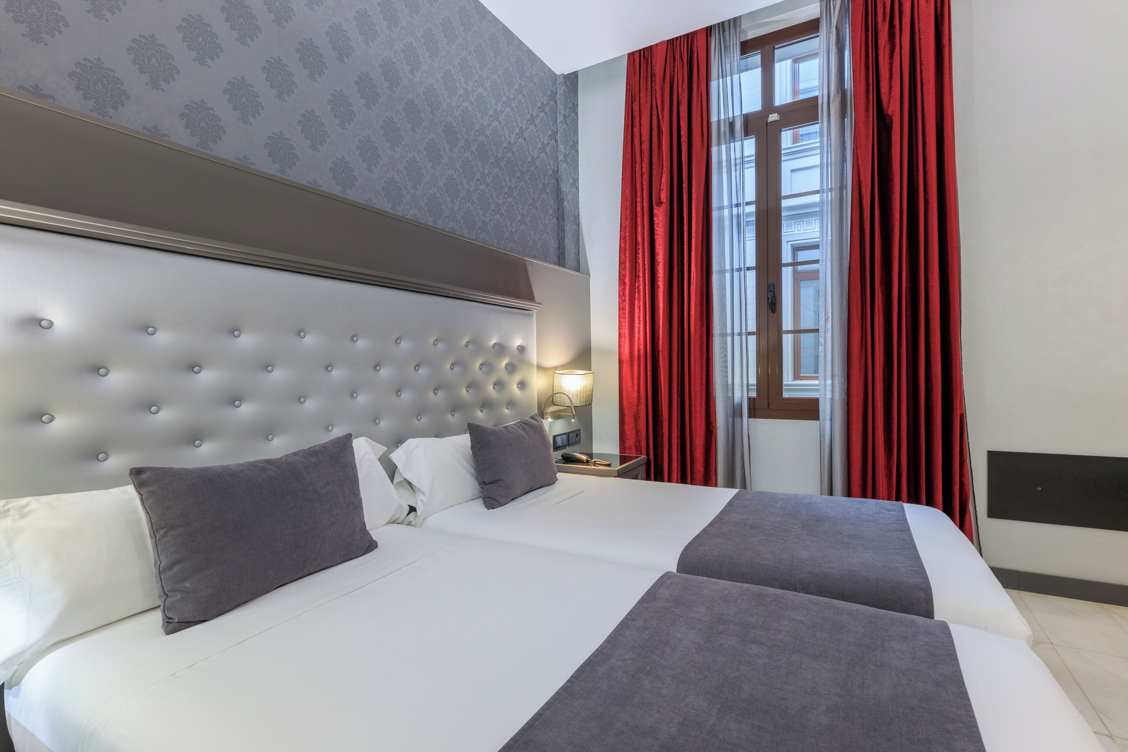 Habitación de hotel con una cama grande, cabecera, mesita de noche y ventana con cortinas rojas.
