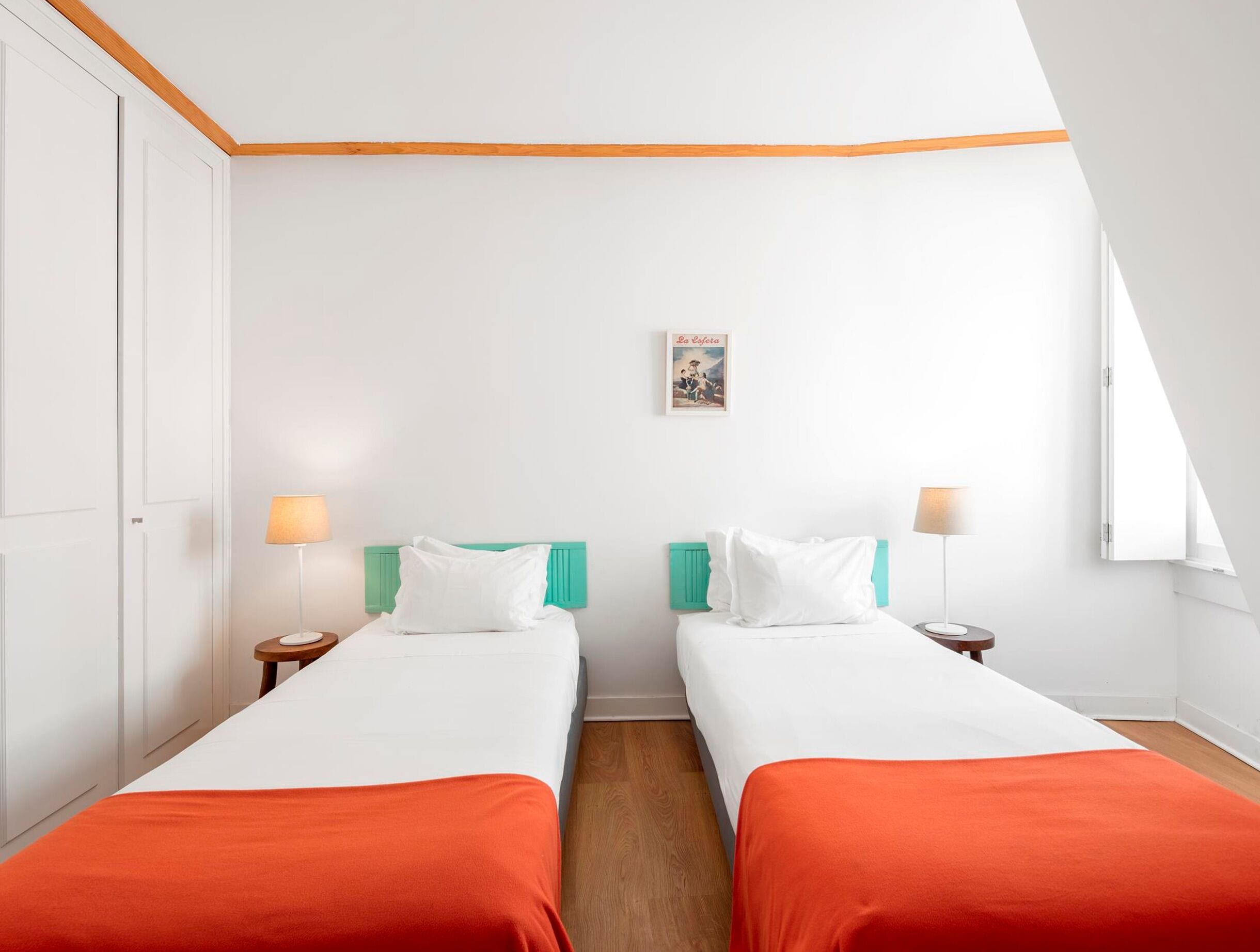A hotel room with two beds, white bedding, orange throw blankets, teal headboards, and a small table with a lamp.