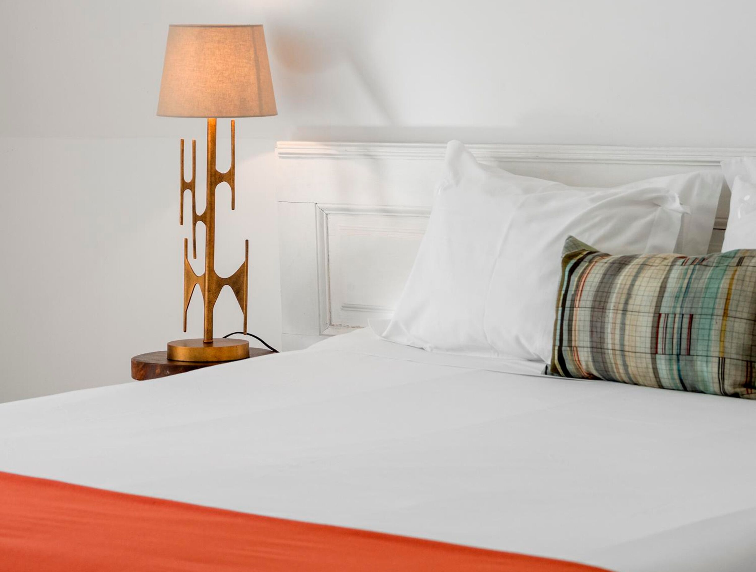 A bed with white bedding, a plaid pillow, a wooden lamp with a unique design, and a white headboard.