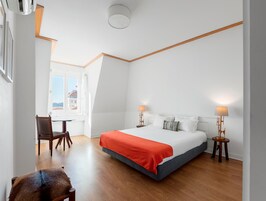 Quarto de hotel com cama, duas luminárias de cabeceira, uma cadeira, uma mesinha e janela com vista.