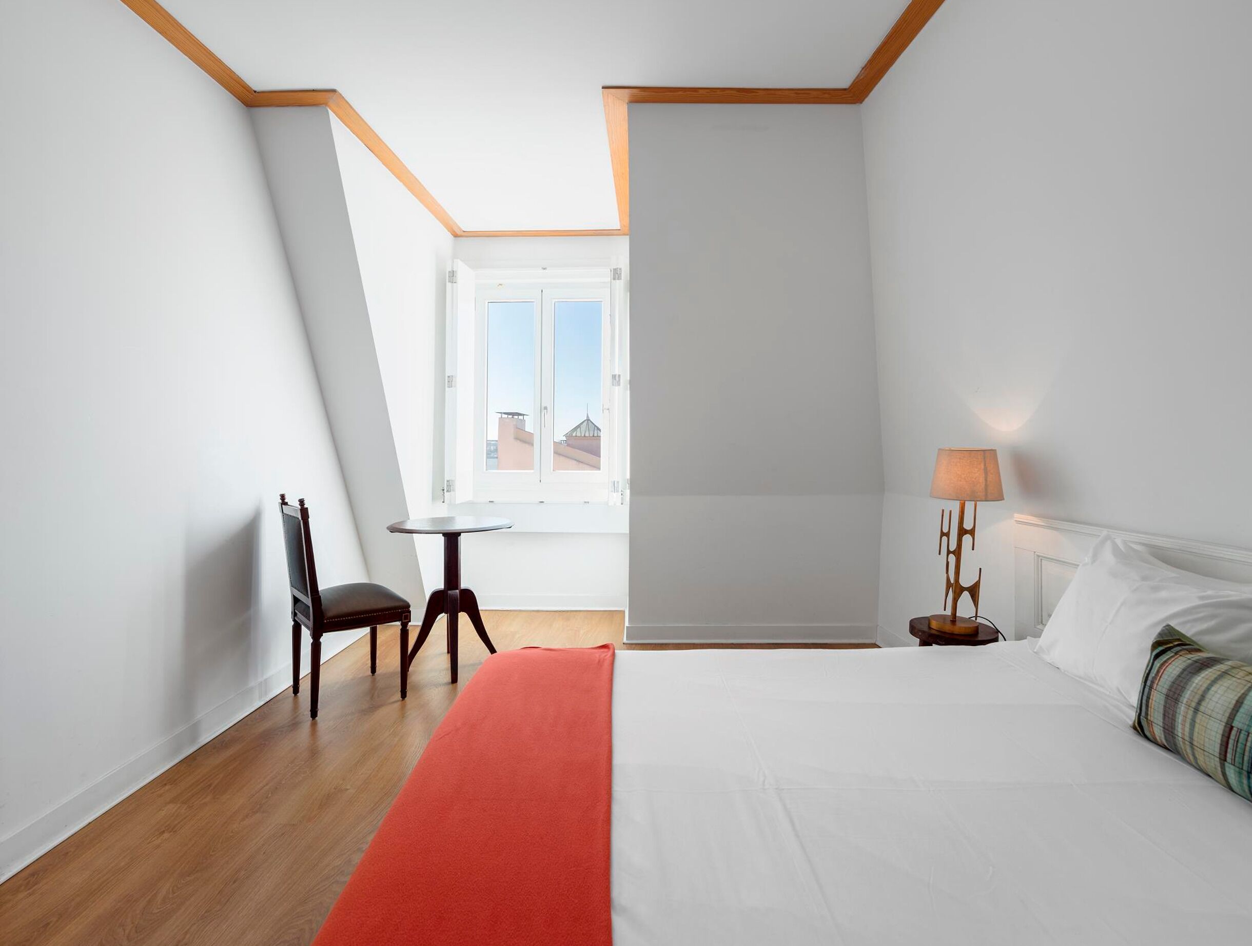 A hotel room with a bed, a chair, a small table, a lamp, and a window with a view.