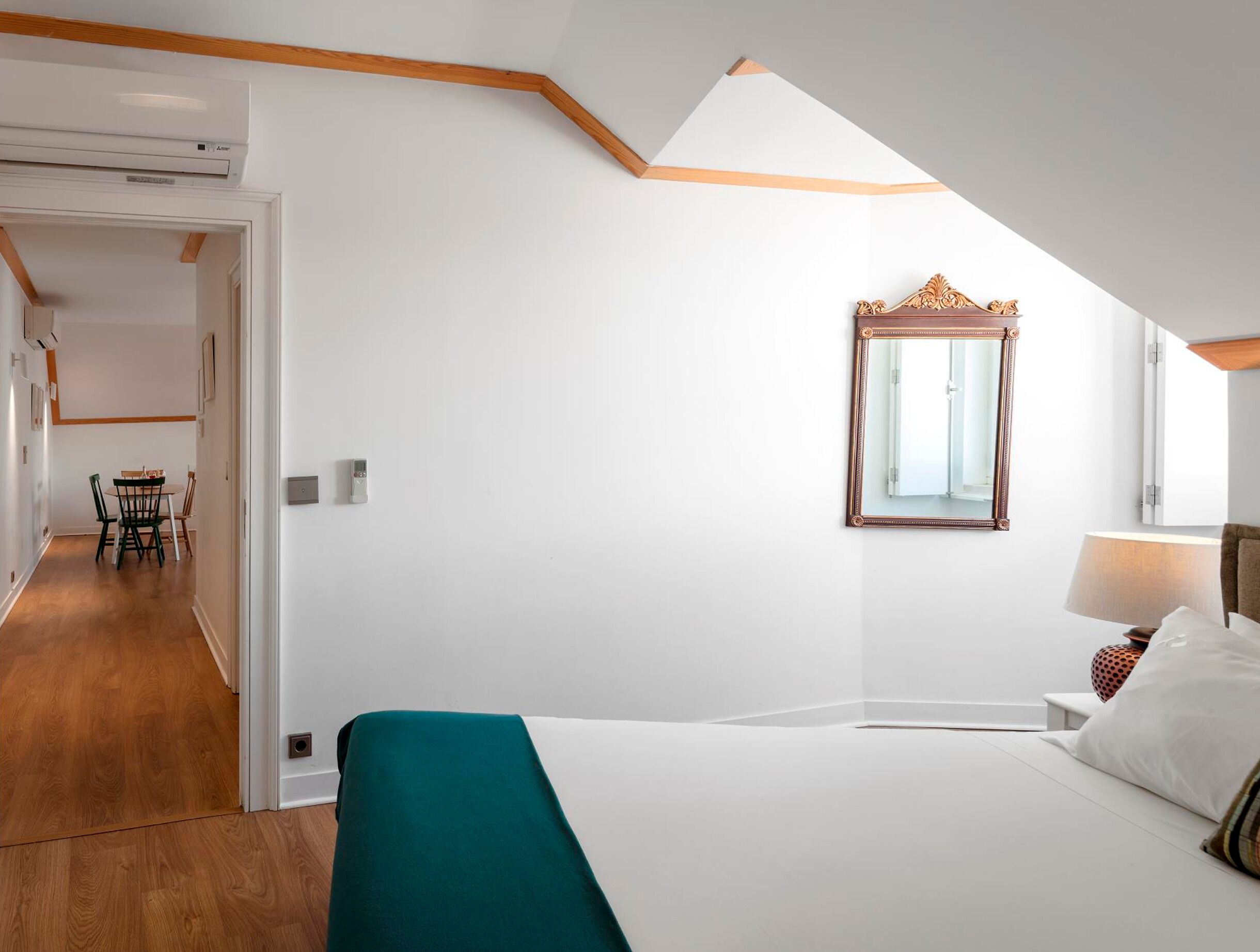 A bedroom with a bed, a mirror, a bedside table, a lamp, and a view of a hallway with a dining table and chairs.