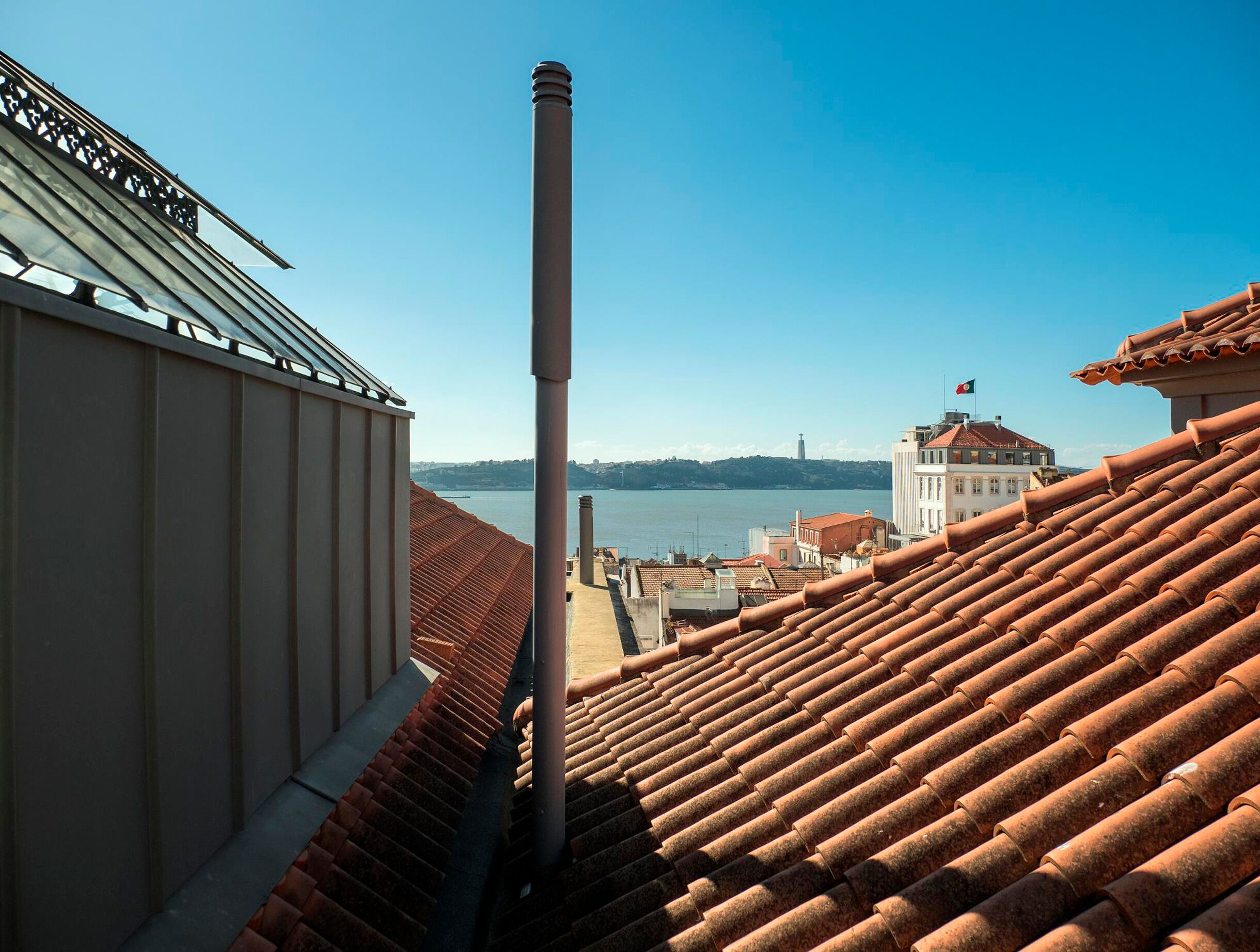 Vista de un edificio con techo de metal y chimenea, con vista a una masa de agua y un paisaje urbano distante.