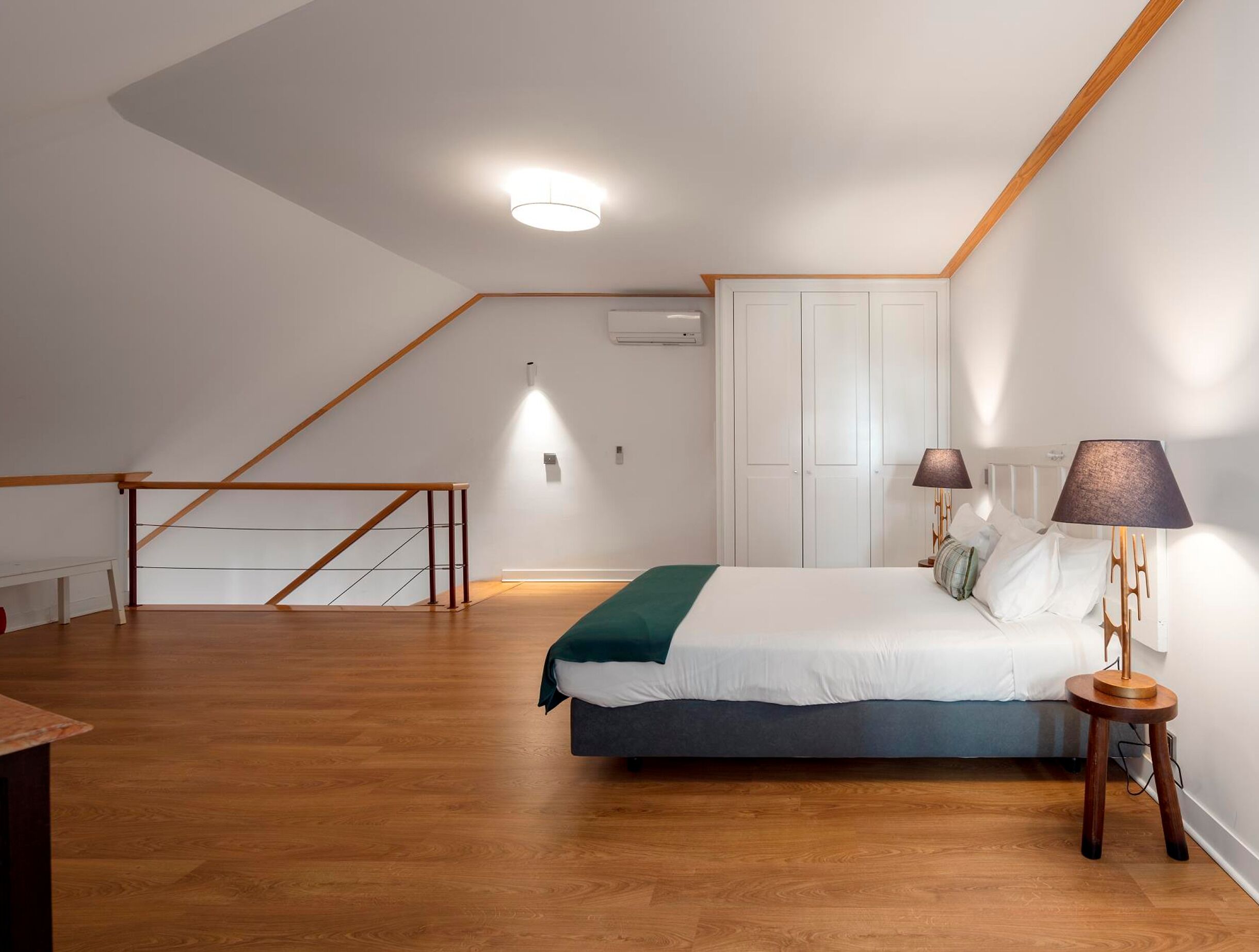 Un dormitorio con cama, lámparas de noche, suelo de madera, un armario blanco y una escalera.