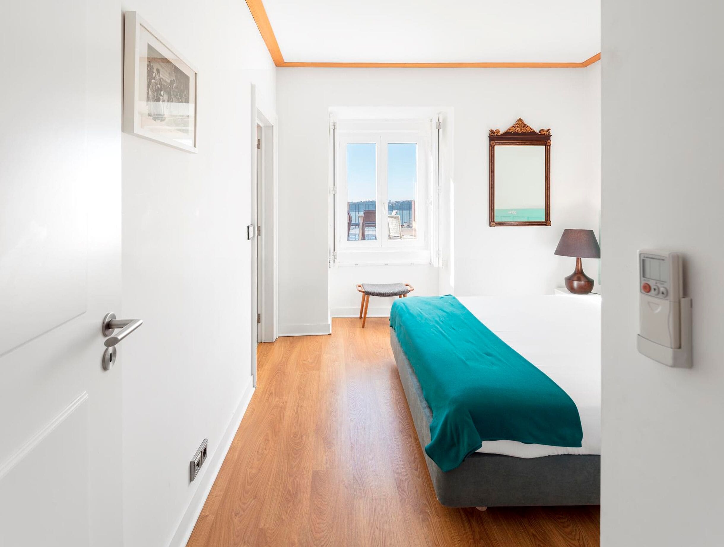 A bedroom with a bed, a window with a view, a bedside table with a lamp, and a thermostat on the wall.