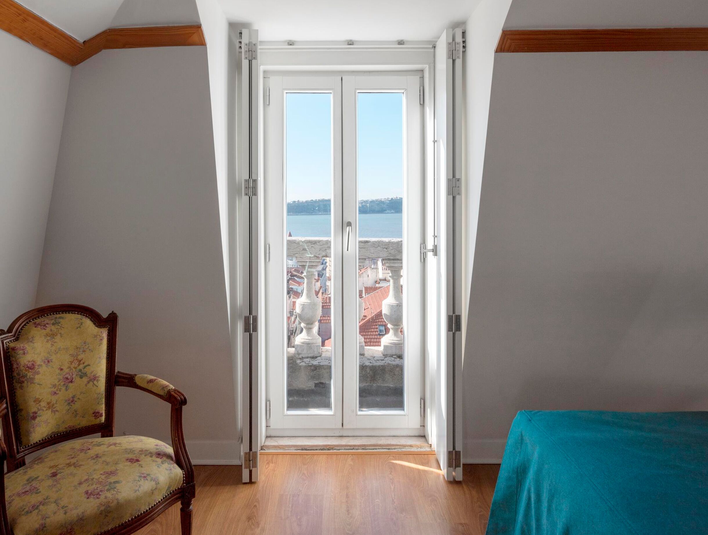 Una habitación con una silla de madera, una mesa turquesa y un ventanal corredero que ofrece vistas al agua y edificios.