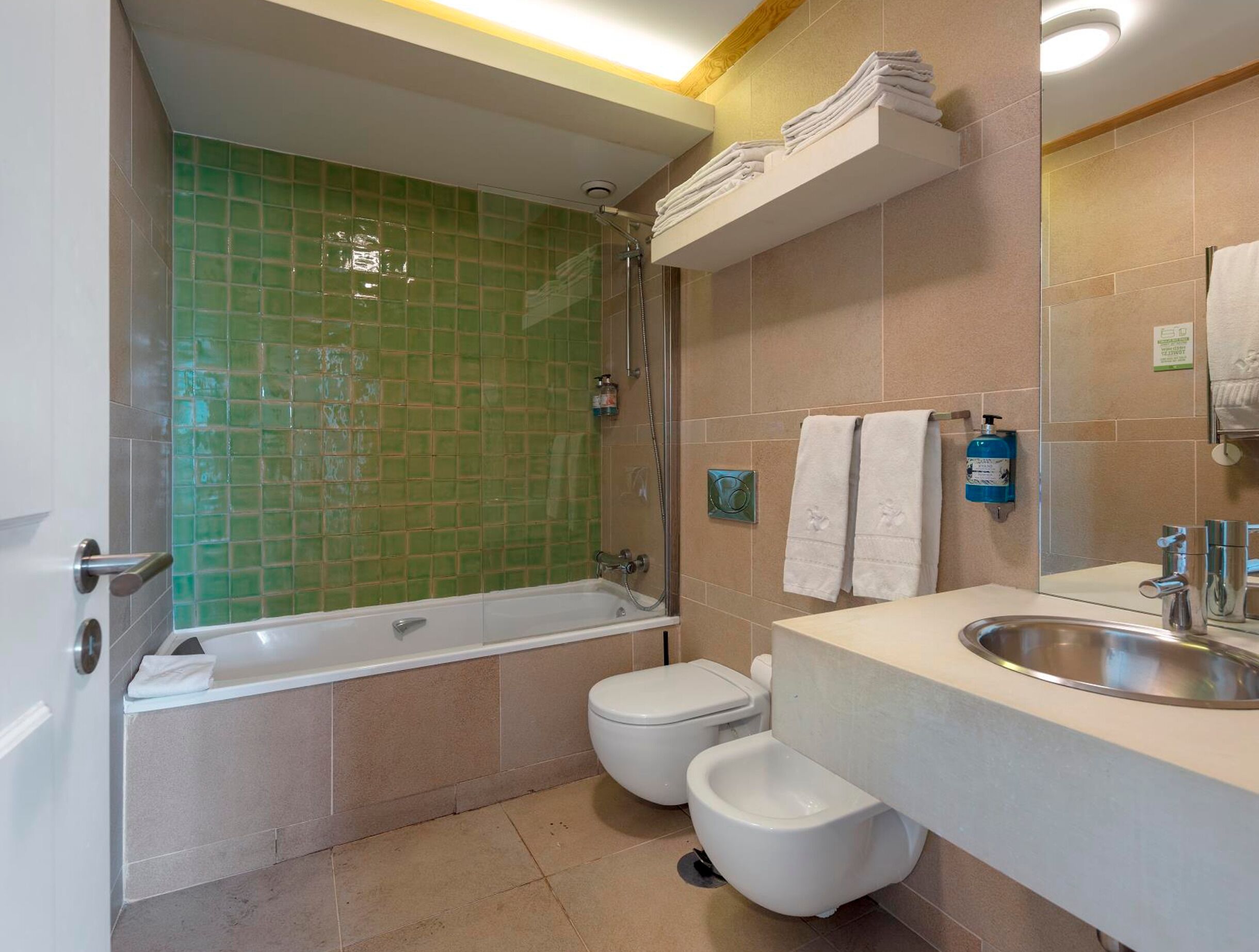 Un baño con ducha revestida de azulejos verdes, una bañera blanca, inodoro y lavamanos con espejo.