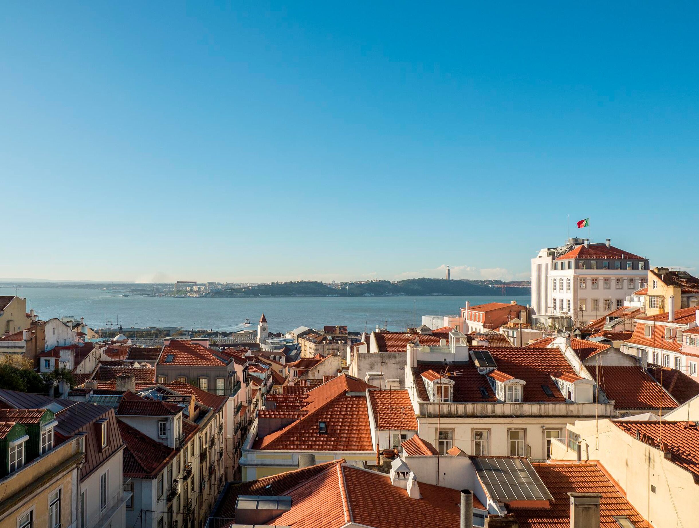 Un paisaje urbano costero con edificios de tejados de terracota, un edificio blanco destacado con una bandera, y una vista despejada del mar.