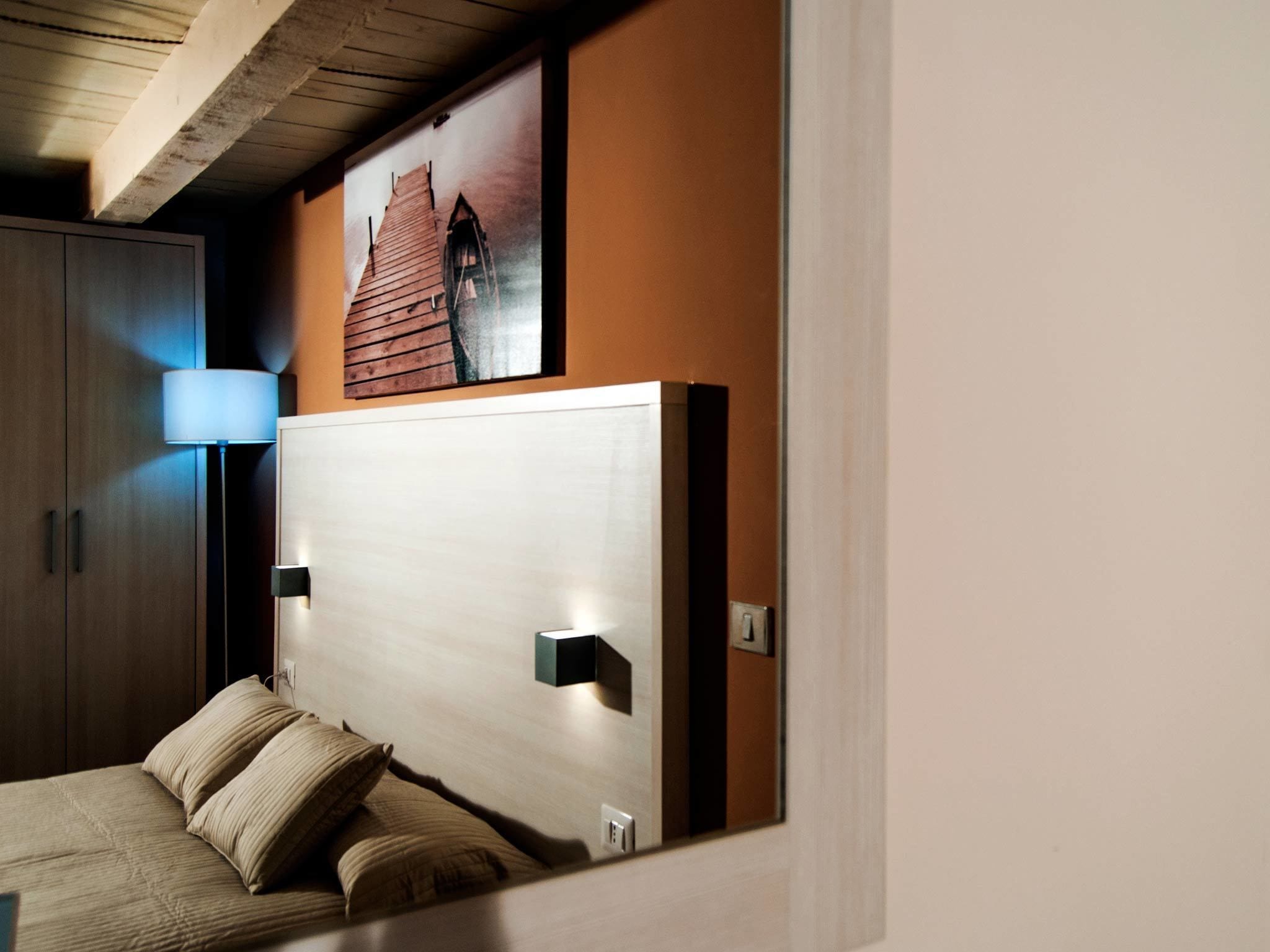 A modern bedroom with a wooden headboard, a bed with beige bedding, a nightstand, a wall-mounted lamp, and a framed picture above the headboard.