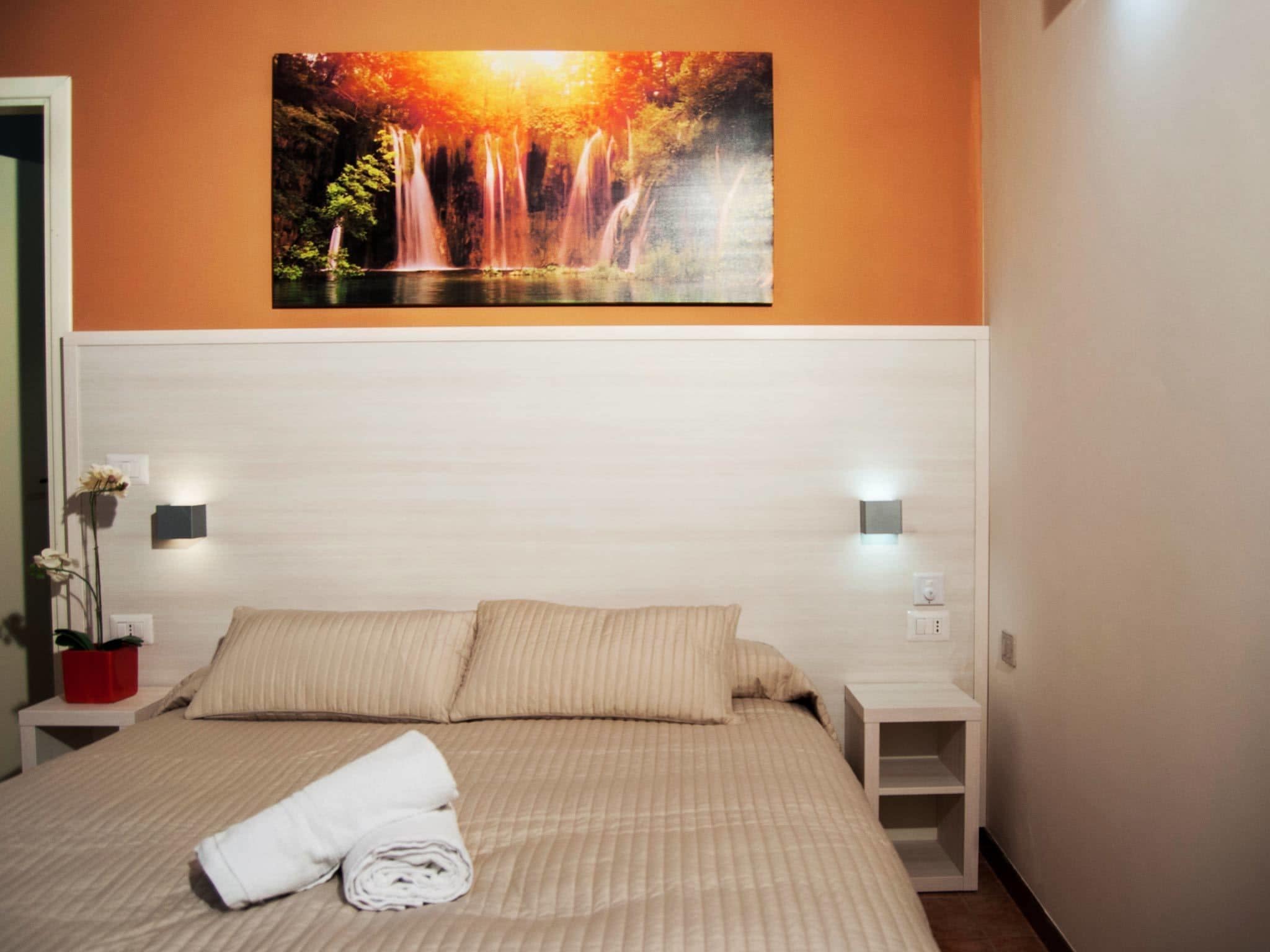 A neatly made bed with a folded towel, a painting of a waterfall, and two bedside tables with wall-mounted lights.
