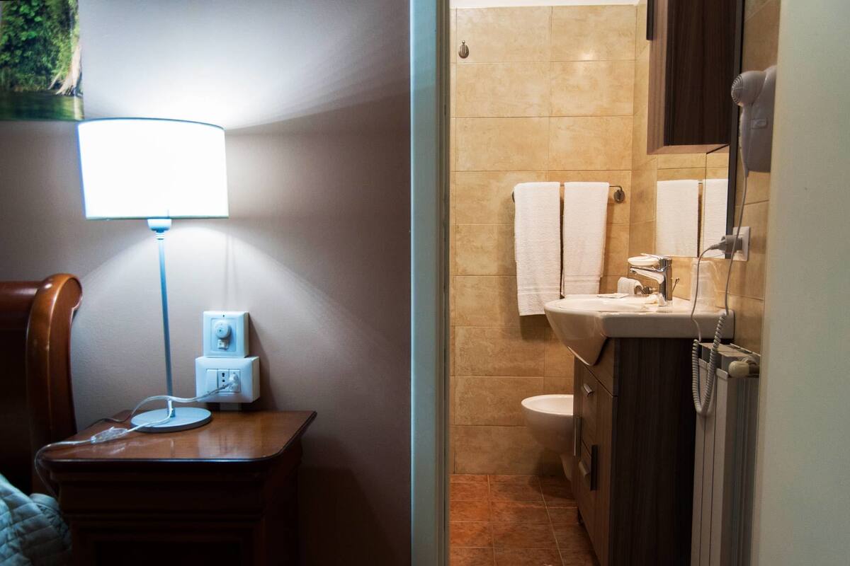 Un baño con lavamanos, inodoro y perchero. Hay una mesa con lámpara y toma de corriente.