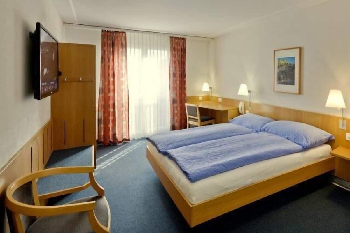Habitación de hotel con una cama doble, un escritorio con silla, un televisor y una ventana con cortinas.