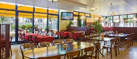Un restaurante espacioso con varias mesas y sillas, ventanas amplias y una pantalla de televisión.