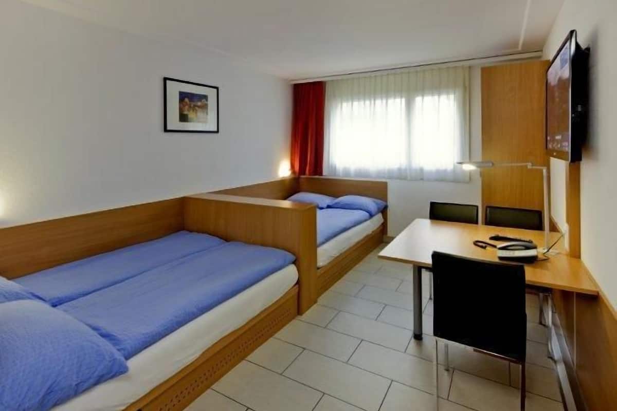 Una habitación de hotel pequeña con dos camas individuales, un escritorio con silla, un televisor montado en la pared y una ventana con cortinas.