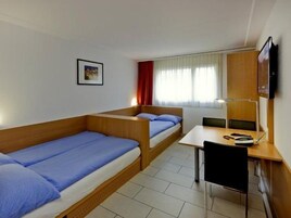 Una habitación de hotel pequeña con dos camas individuales, un escritorio con silla, un televisor montado en la pared y una ventana con cortinas.