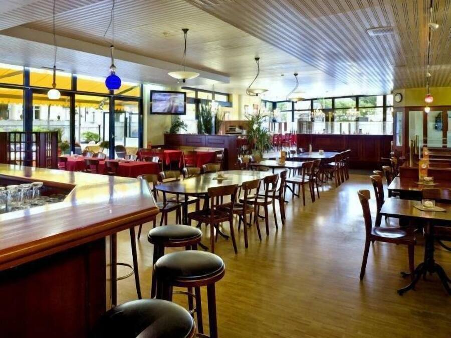 A restaurant with wooden tables and chairs, a bar area, and large windows allowing natural light.