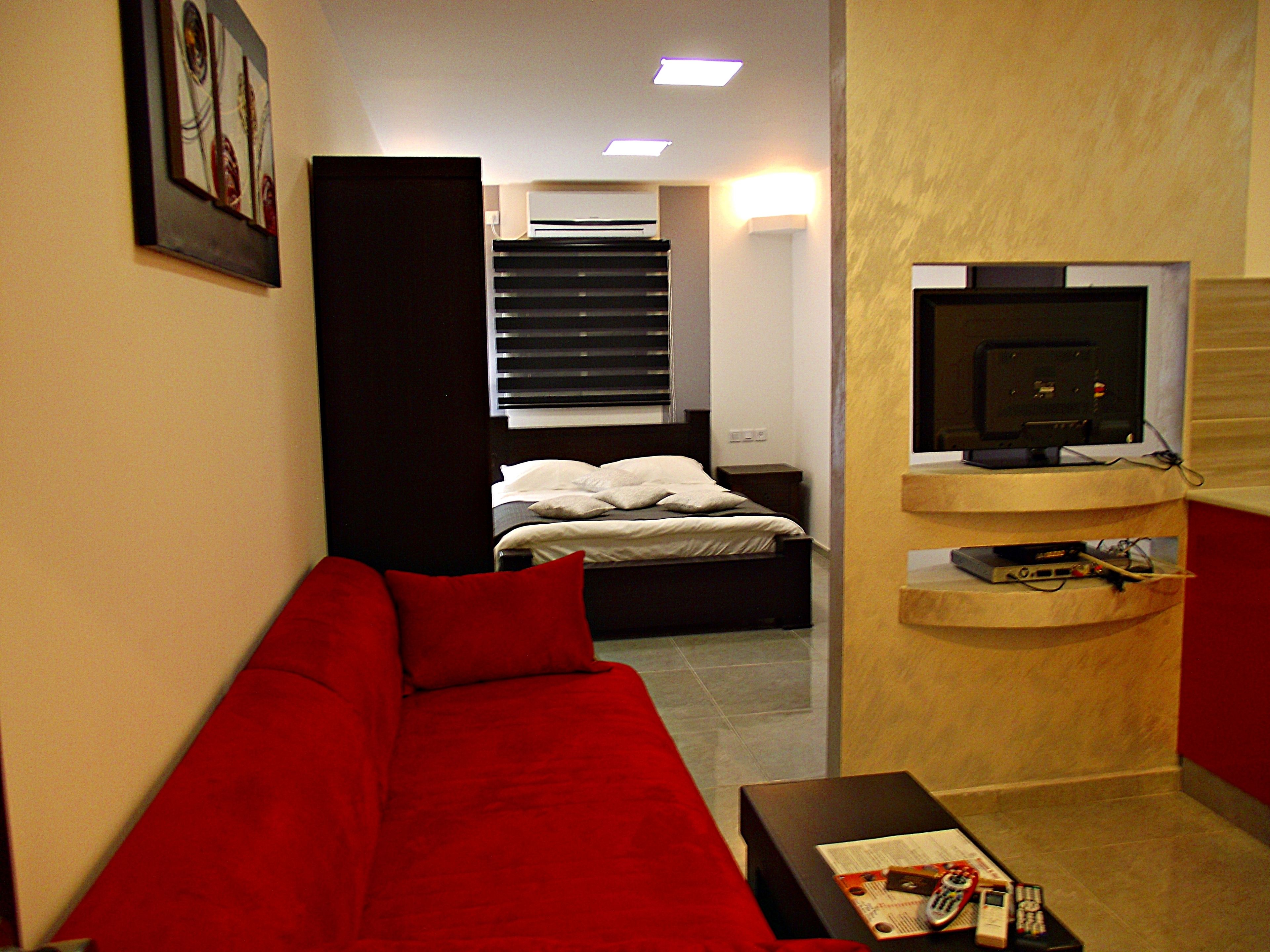 Habitació senzilla amb un sofà vermell, un llit amb roba de llit blanca, un televisor sobre un suport de fusta i una taula petita amb dispositius electrònics.