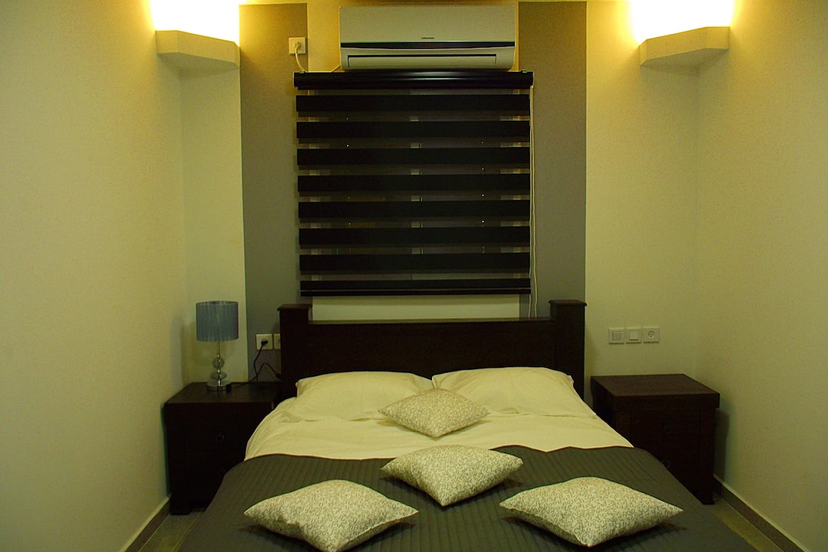 A hotel room with a bed, bedside tables, a lamp, and an air conditioner.
