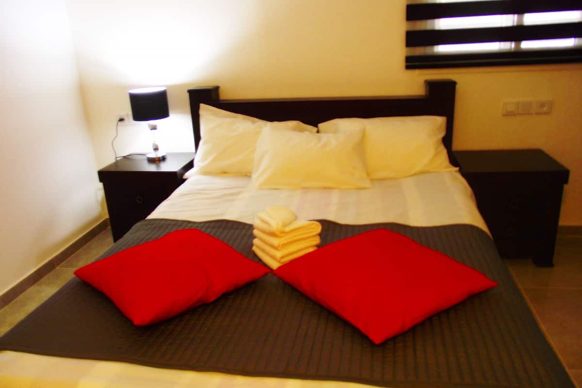 A neatly made bed with white and red pillows, a folded towel, and a bedside table with a lamp.