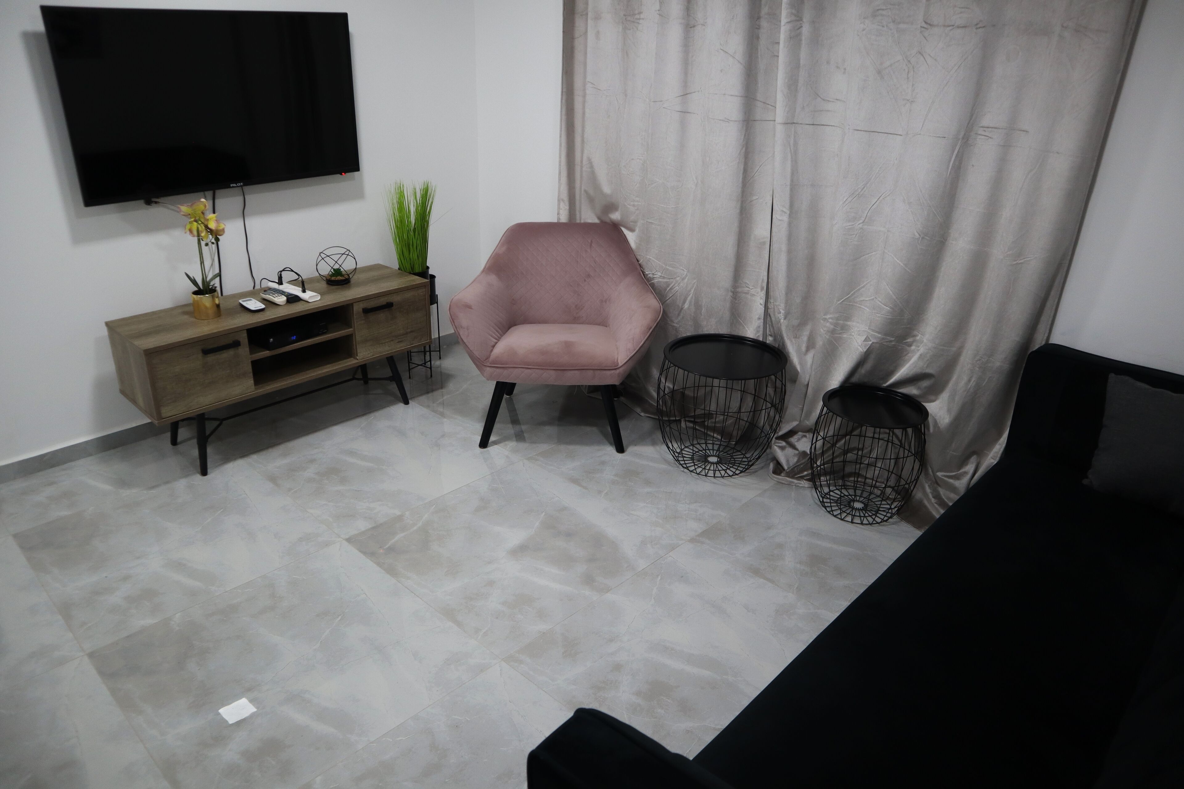 A living room with a flat-screen TV mounted on the wall, a wooden console table with decorative items, a pink armchair, and a black sofa.