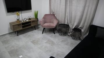 A living room with a flat-screen TV mounted on the wall, a wooden console table with decorative items, a pink armchair, and a black sofa.