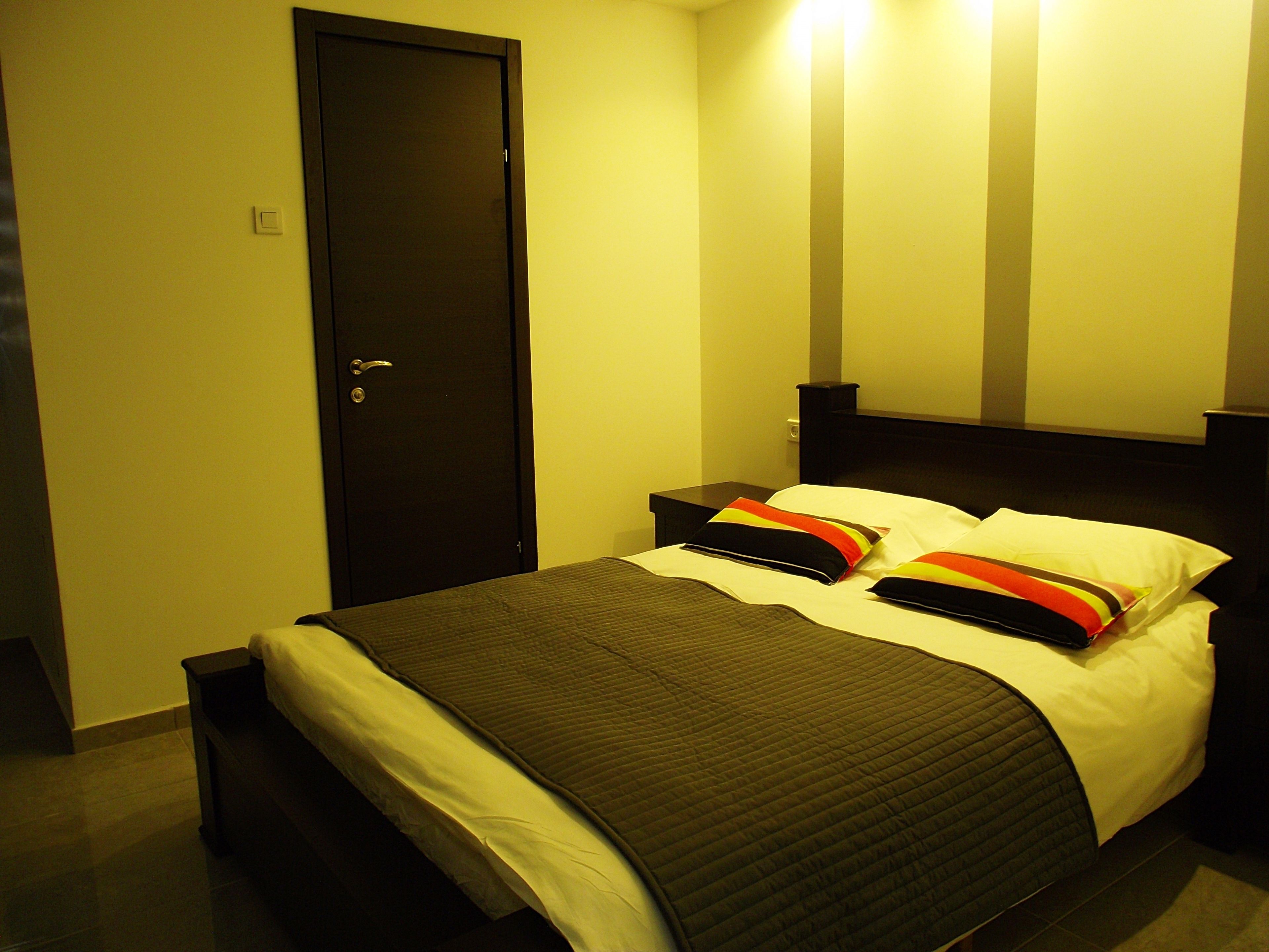 Una camera d'albergo con un letto, un comodino, una porta e una lampada a parete.