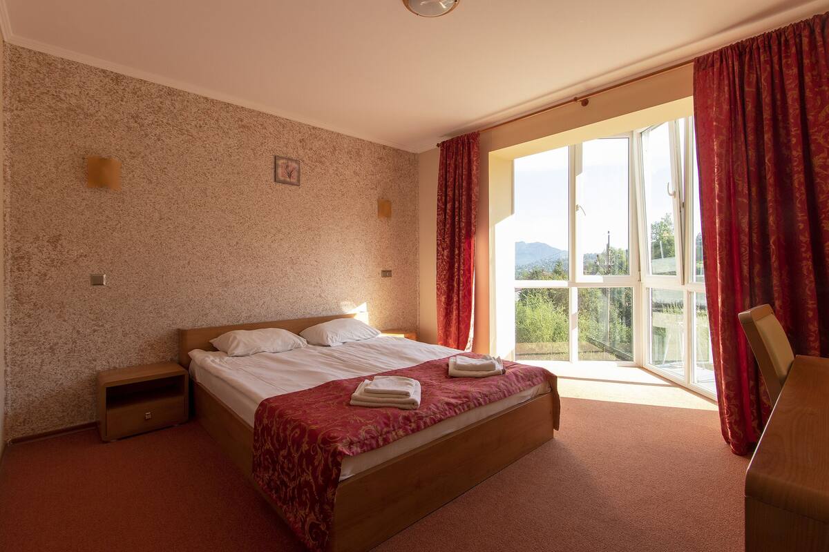 A bedroom with a bed, a nightstand, a window with curtains, and a view of greenery outside.