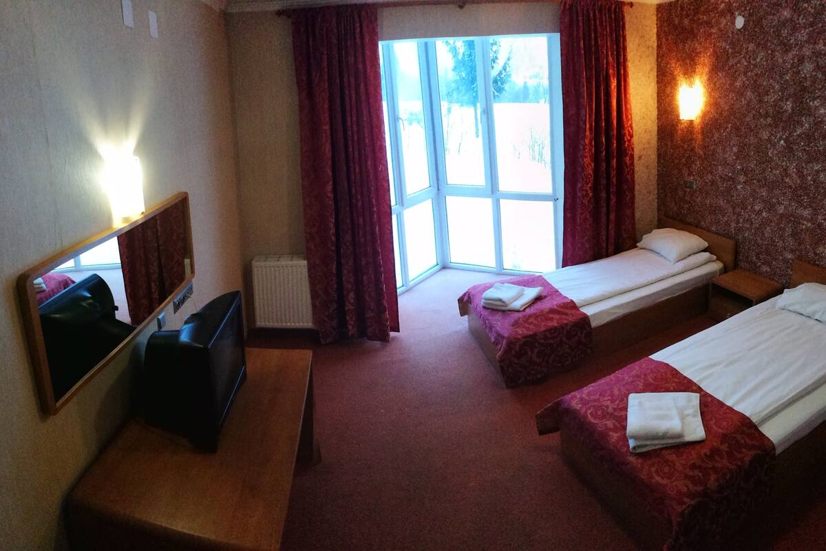 A hotel room with two single beds, a TV, a radiator, and a window with curtains.