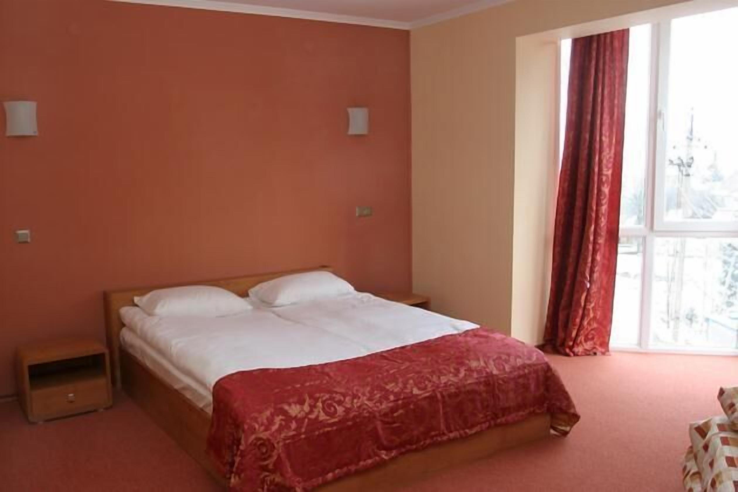 A bedroom with a bed, a nightstand, a window with curtains, and a view of a building outside.