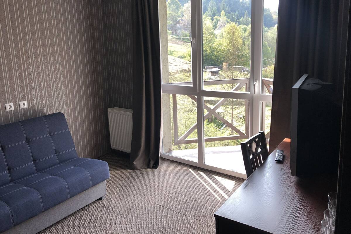 A room with a sofa, a table, a radiator, and a large window with a view of greenery and buildings.