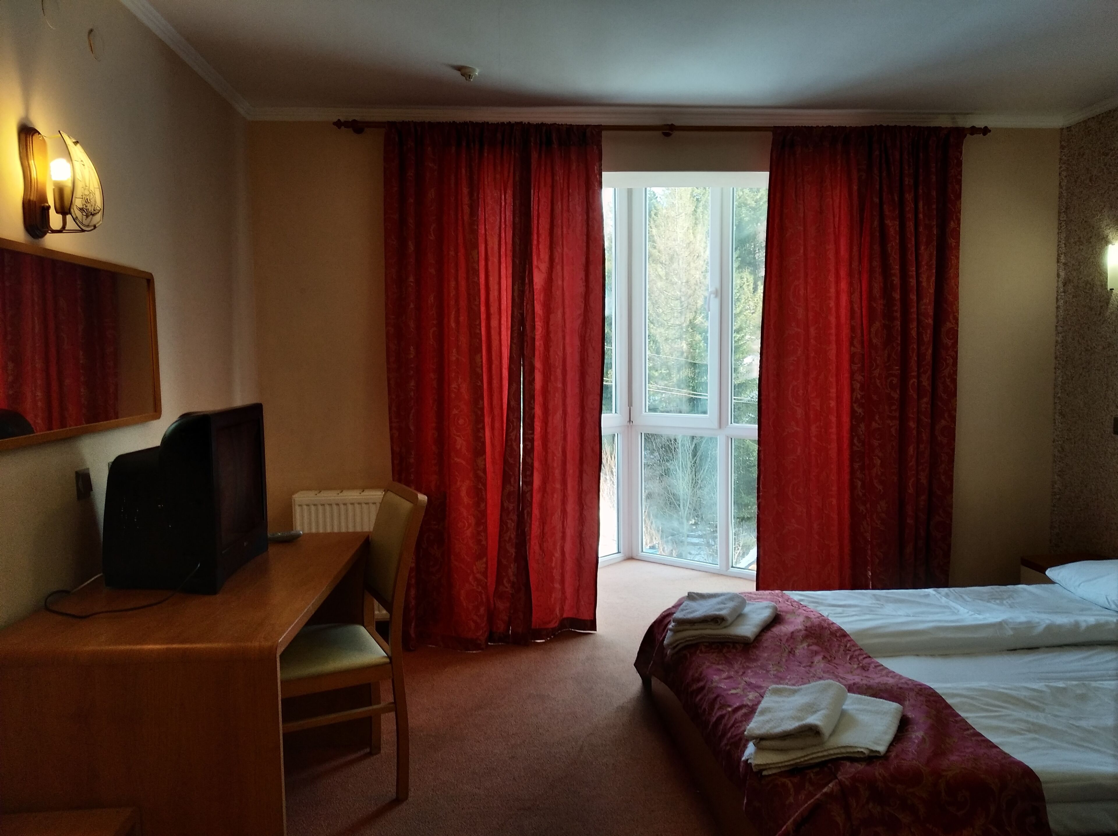A hotel room with a bed, a desk with a chair, a TV, and a window with red curtains.