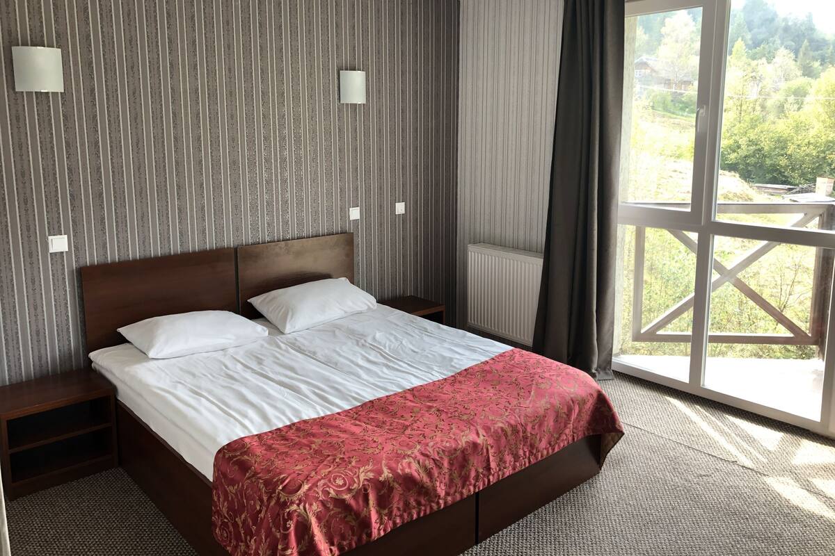 A hotel room with a large bed, two bedside tables, a radiator, and a view of greenery through a window.
