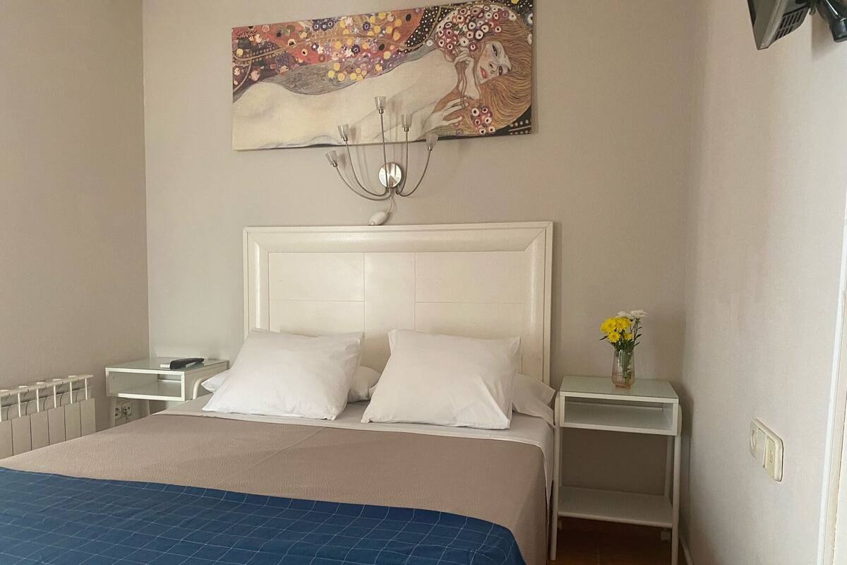 A bedroom with a large bed, white headboard, two bedside tables, a nightstand, a wall-mounted lamp, and a painting above the bed.