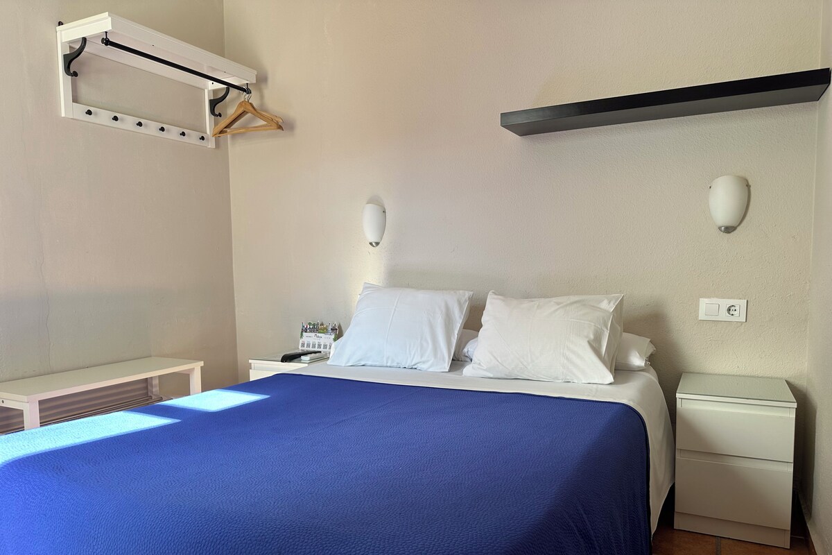 A neatly arranged bedroom with a bed, bedside tables, a nightstand, a wall-mounted shelf, and a wall socket.