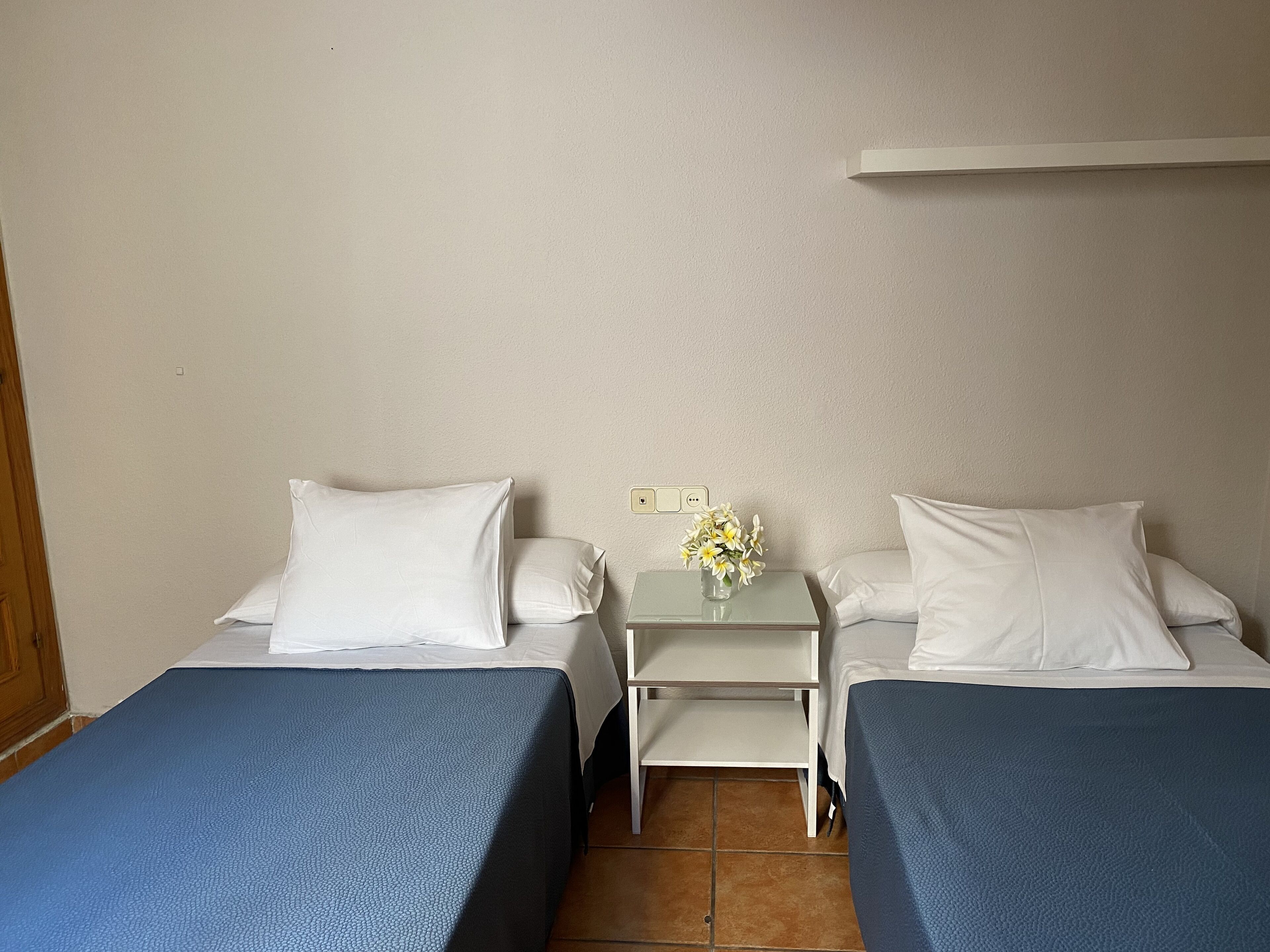 Una habitación de hotel con dos camas individuales, una mesita con un jarrón de flores y un estante en la pared.