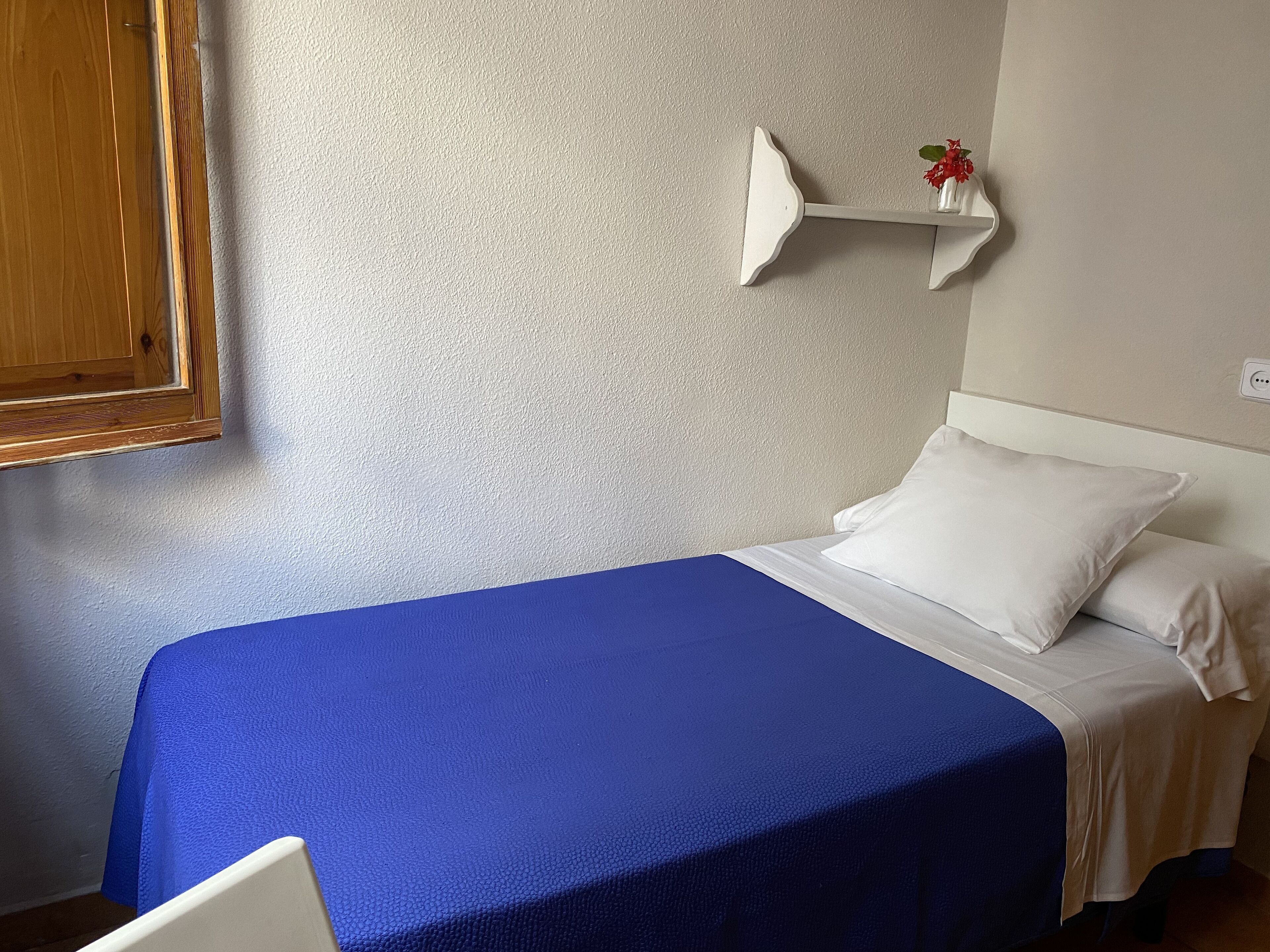 A single bed with a blue bedspread, white pillows, and a white headboard. There is a white shelf with a red flower on the wall to the right of the bed.
