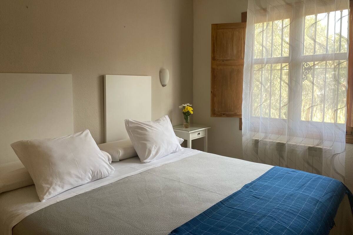 A neatly made bed with white bedding and a blue accent, a small bedside table with a vase of flowers, and a window with sheer curtains.