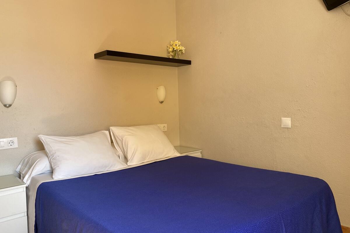 A bedroom with a bed, white pillows, a blue bedspread, a nightstand, a wall-mounted shelf with a vase of flowers, and a wall socket.