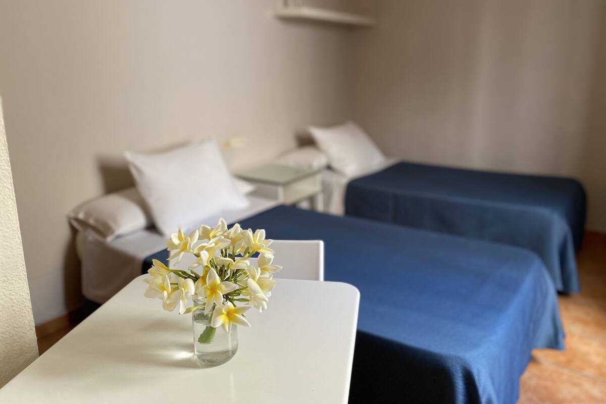 A hotel room with two beds, a small table with a vase of flowers, and a white wall.