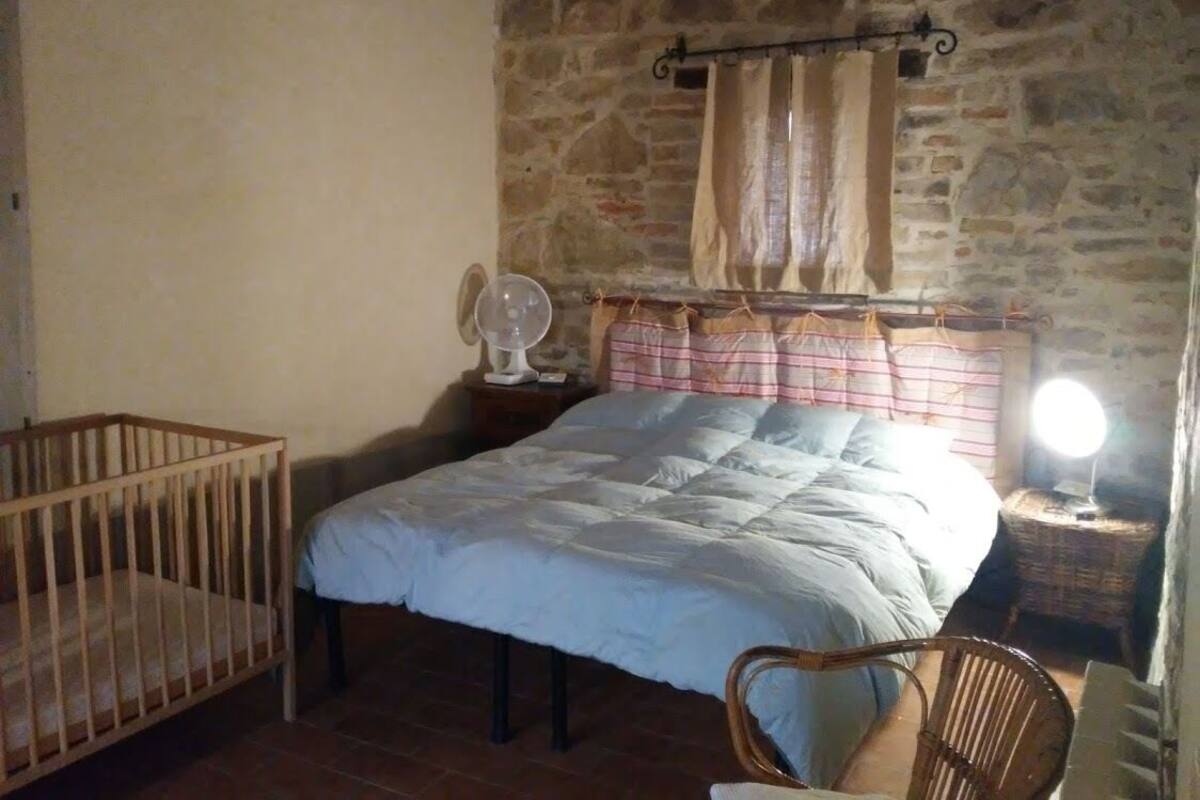 Een slaapkamer met een stenen muur, een bed met een blauwe quilt, een houten kinderbedje, een nachtkastje met een lamp en een rieten stoel.