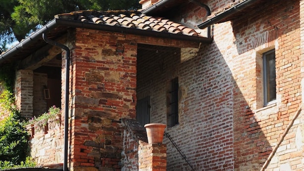Um edifício rústico de pedra com telhado de telhas, chaminé de tijolos e um pequeno alpendre com um vaso de planta.