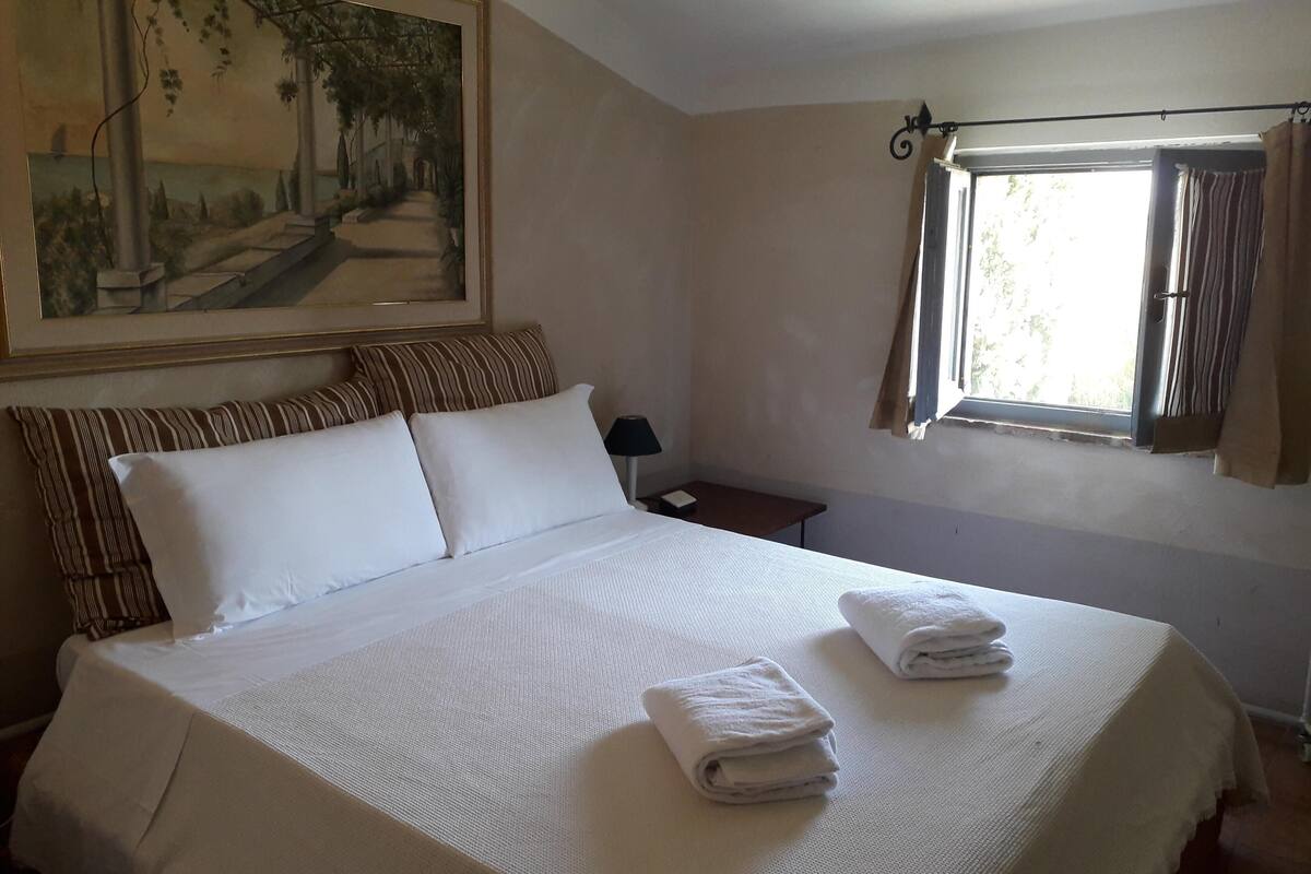 Een keurig opgemaakt bed met witte lakens en twee gevouwen handdoeken, een landschapsschilderij aan de muur, en een raam met gordijnen.