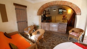 Um interior acolhedor com soalho de pedra, mobiliário de vime e uma área de cozinha visível através de uma porta em arco de tijolos.