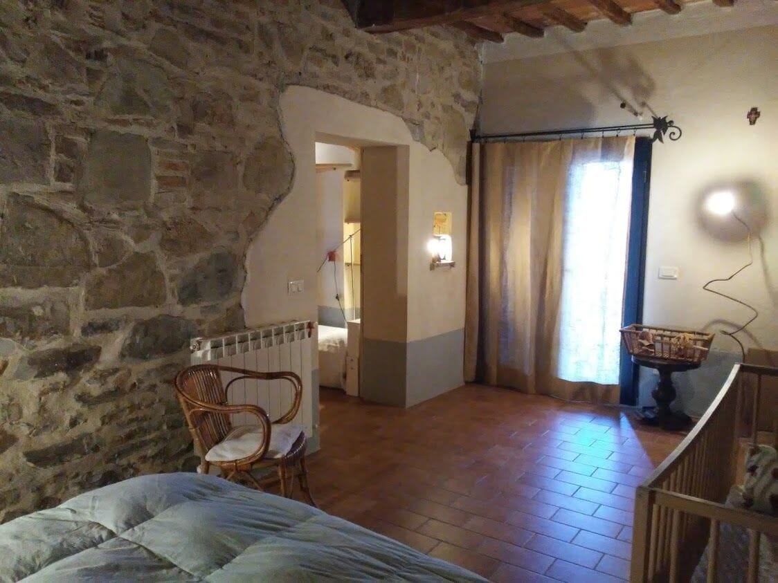 A room with a stone wall, a bed, a chair, a radiator, a lamp, and a window with curtains.