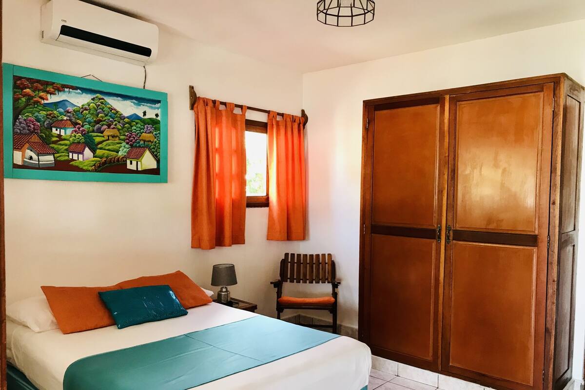 Een slaapkamer met een bed, nachtkastje, stoel, kledingkast en een schilderij aan de muur.
