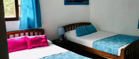 Quarto com duas camas de solteiro, mesa de cabeceira com luminária, pintura na parede e janela com cortinas azuis.