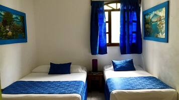 A room with two beds, each with a blue bedspread and pillow, a small nightstand with a lamp, and a window with blue curtains.
