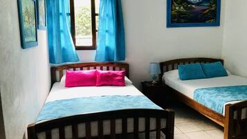 A room with two wooden beds, each with a blue bedspread and pillows, a window with blue curtains, and a painting on the wall.